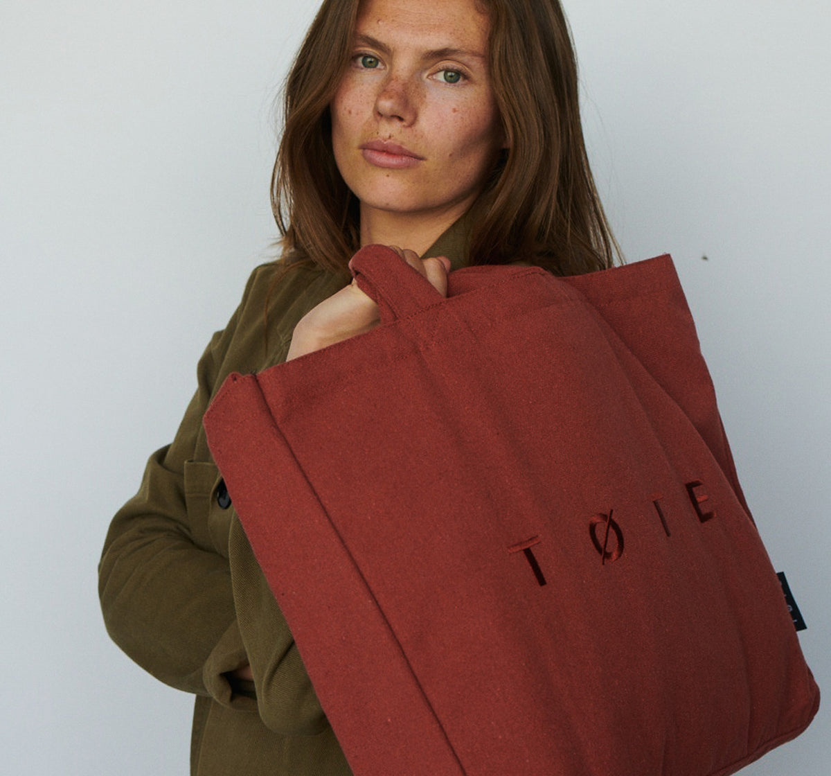 Model carrying a rust TOTE Bag against a light studio wall
